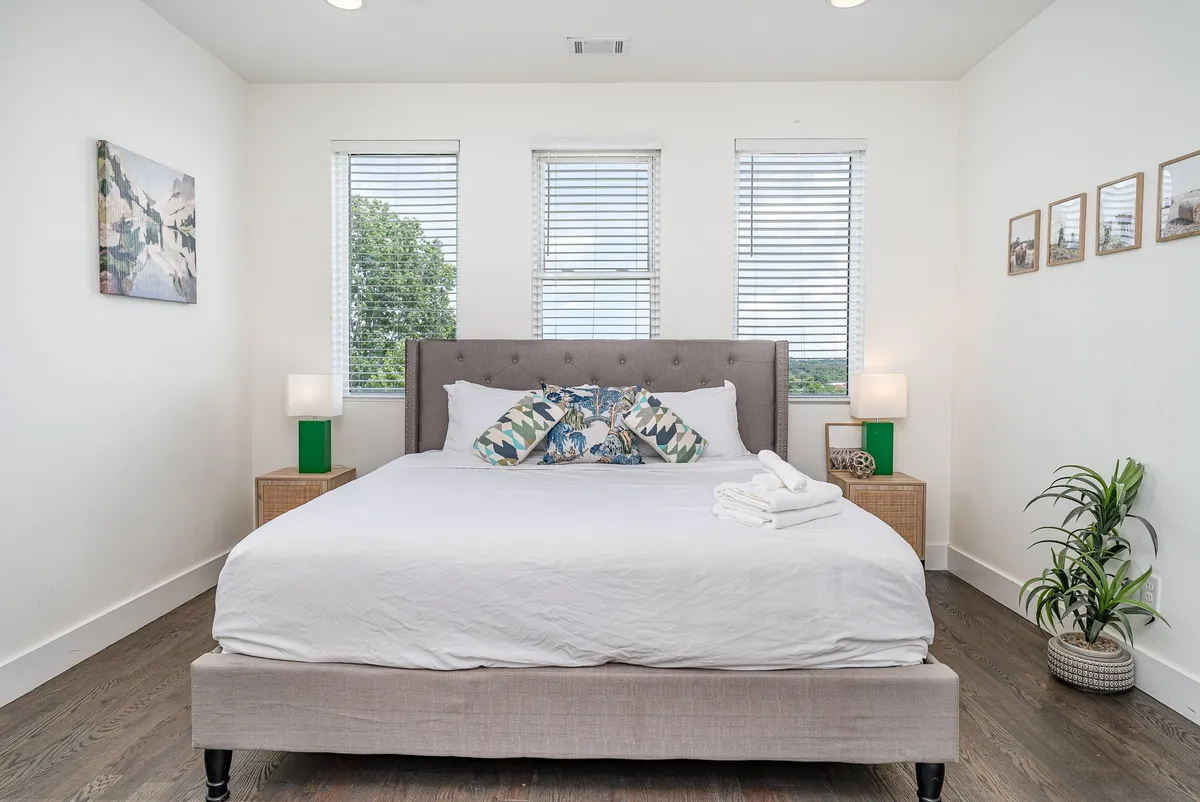 A tranquil bedroom featuring a bed with decorative pillows, nightstands topped with lamps, framed artwork above the headboard, and a lush indoor plant, bathed in natural light. The atmosphere is calm and cozy, evoking a restful, luxurious retreat ideal for a primary suite.
