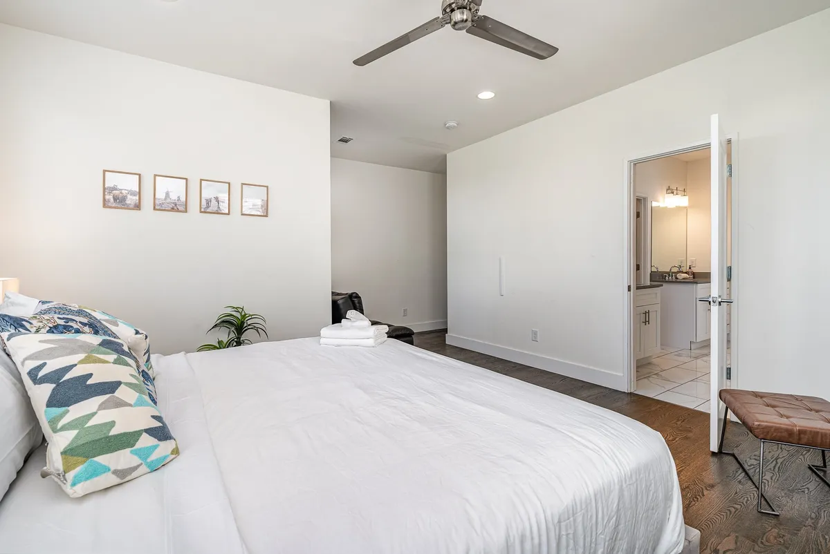 An elegant bedroom with a well-made bed adorned with decorative pillows, stylish framed pictures on the wall, a leafy plant on the side table, and an open door showing a glimpse of an en-suite bathroom. The mood is serene and inviting, emphasizing convenience and relaxation.