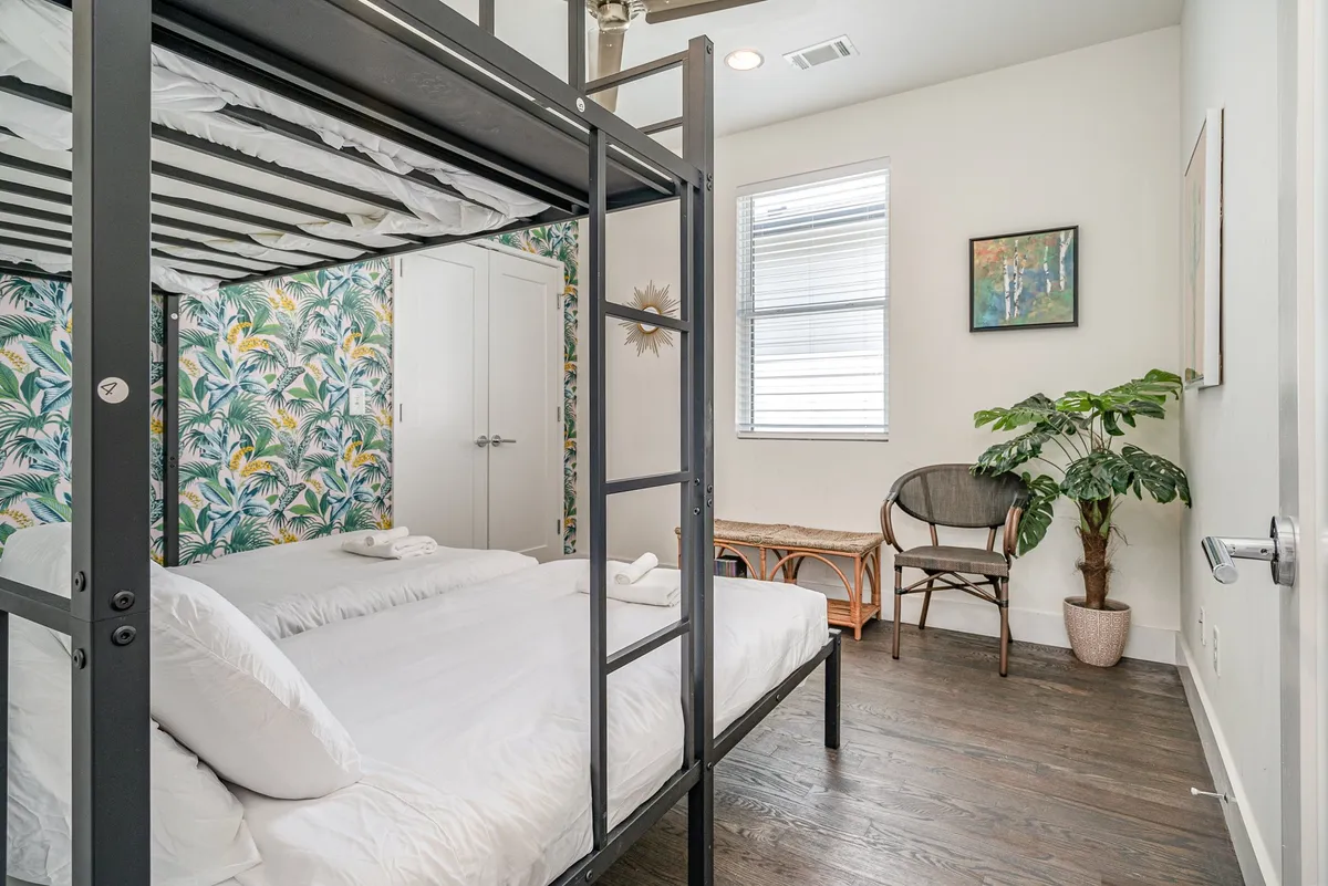 A comfortable and cheerful bedroom with a bunk bed, tropical wallpaper, a potted plant, and a rattan chair. The setup offers a relaxed spot to sip morning coffee or enjoy quiet moments, with decor that feels both fun and soothing.