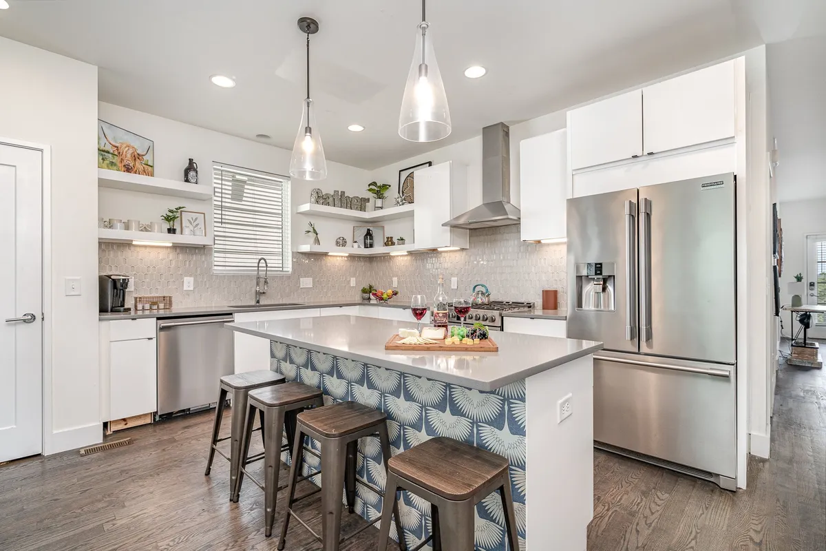 The image showcases a well-equipped kitchen with a prominent island bar, stainless steel appliances, decorative shelving, colorful fruit arrangement, and wine glasses. The casual seating at the island bar encourages both quick breakfasts and evening cocktails—key moments mentioned in the content.