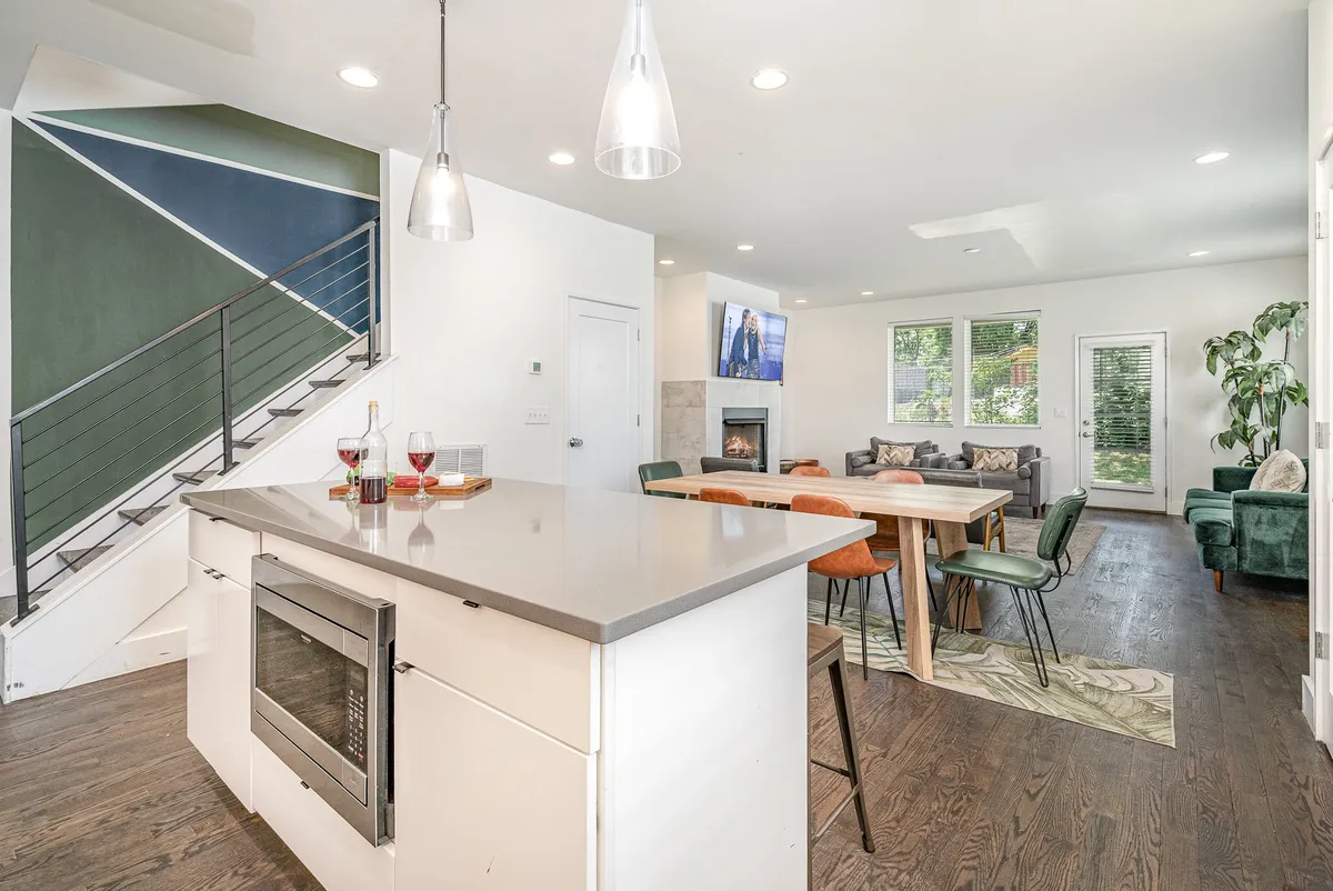 A bright, modern open-plan space featuring a kitchen island, dining area, green staircase, fireplace, and large windows. The setting is inviting and well-equipped, ideal for both everyday living and special occasions.