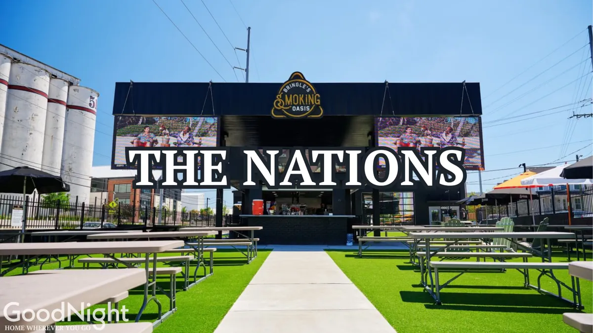 An exterior view of a lively outdoor seating area with large signage and a bustling barbecue food stall. People are socializing and dining, conveying a sense of community and festivity.