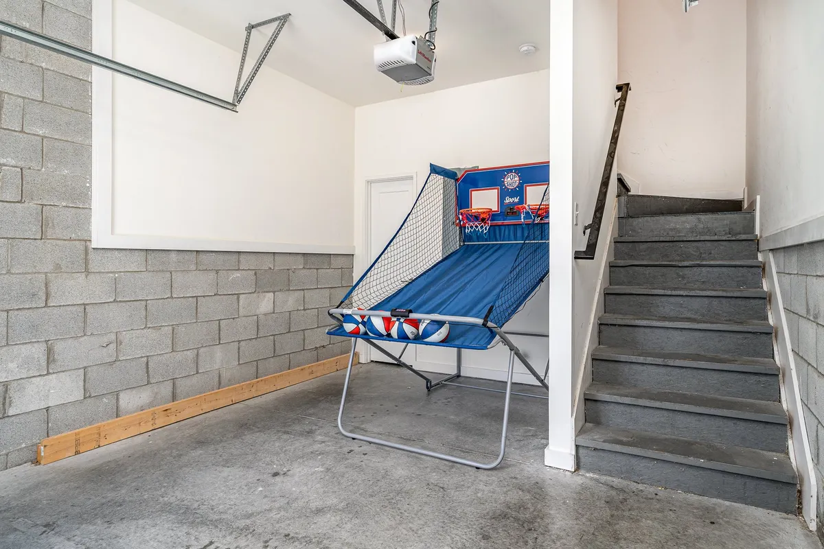 A lively garage scene featuring a basketball hoop game positioned against one wall, with a staircase and garage walls visible in the background. The space feels open and casual, ready for friendly competitions or casual entertainment.