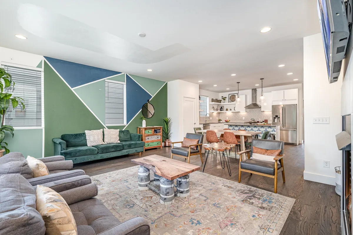 The hero image features a spacious and inviting living area with a bold green accent wall adorned by a modern geometric design. At the center is a stylish wooden coffee table surrounded by plush, comfortable seating—ideal for socializing, relaxing, or enjoying a cozy movie night. The space is bright and contemporary, perfect for gatherings with family and friends.