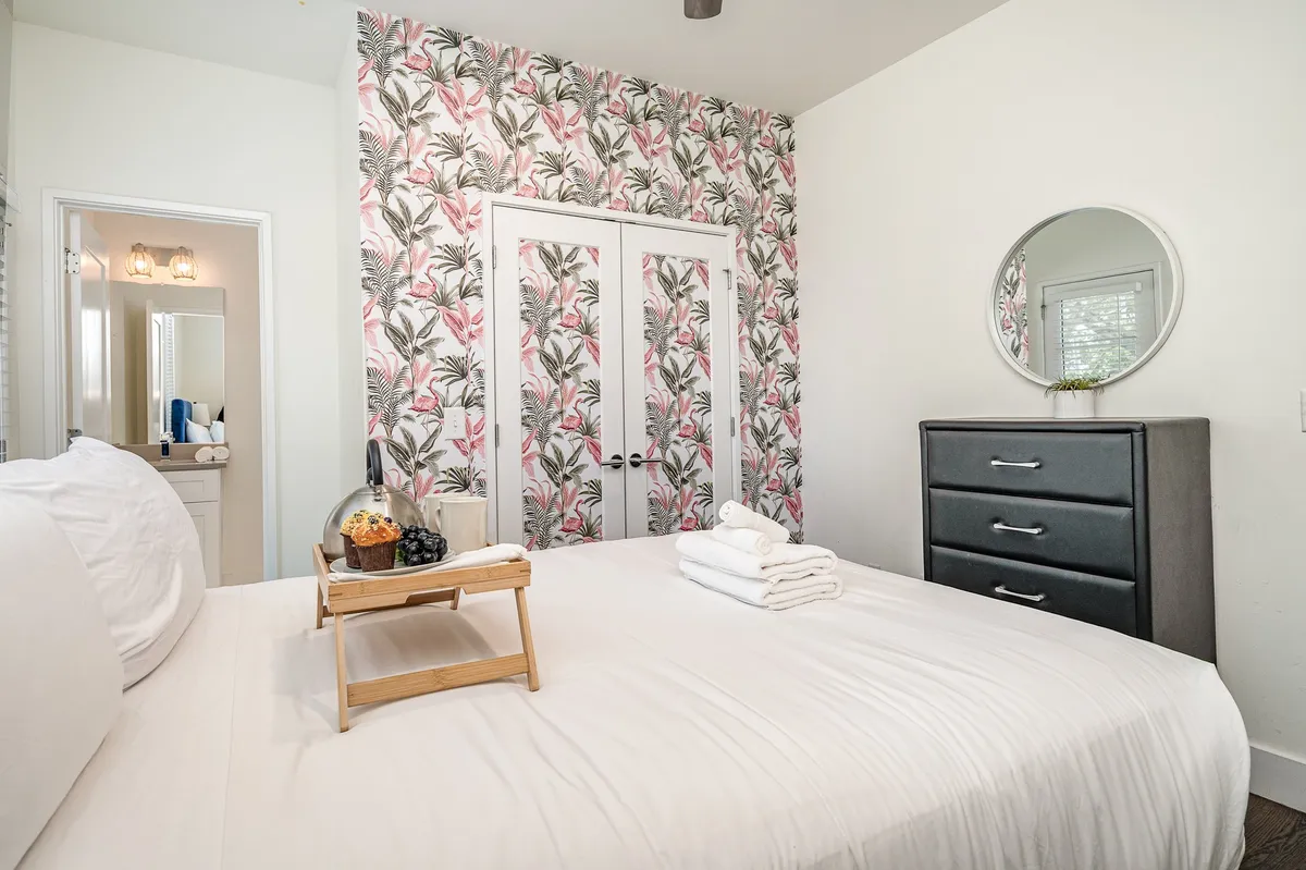 A cheerful queen suite with white linens, tropical patterned wallpaper, a dressing table with a mirror, a chest of drawers, and a breakfast tray with fruit. Sunlight streams through, and the overall effect is refreshing and cozy—perfect for a restful escape with balcony access.