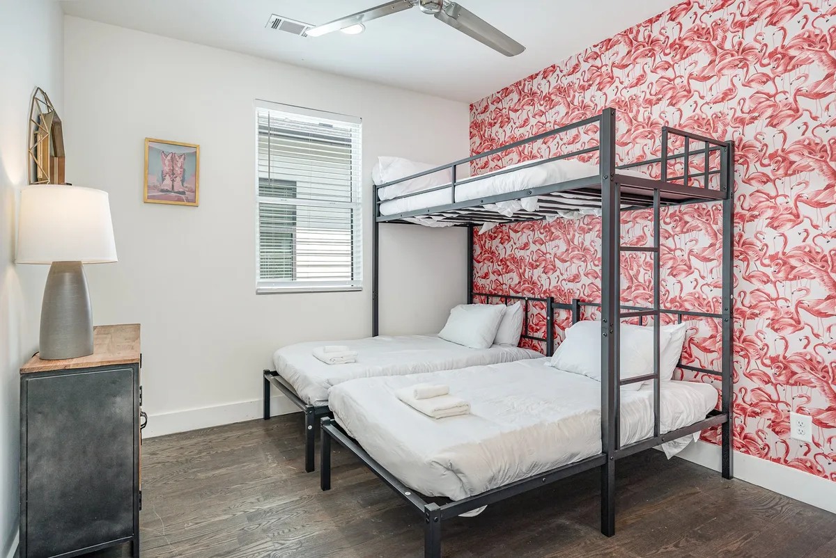 A playful and cozy bunk room with a bunk bed, whimsical flamingo wallpaper, and a nightstand with a lamp. Framed artwork and soft lighting create an inviting atmosphere perfect for relaxing or unwinding after a day in the city.